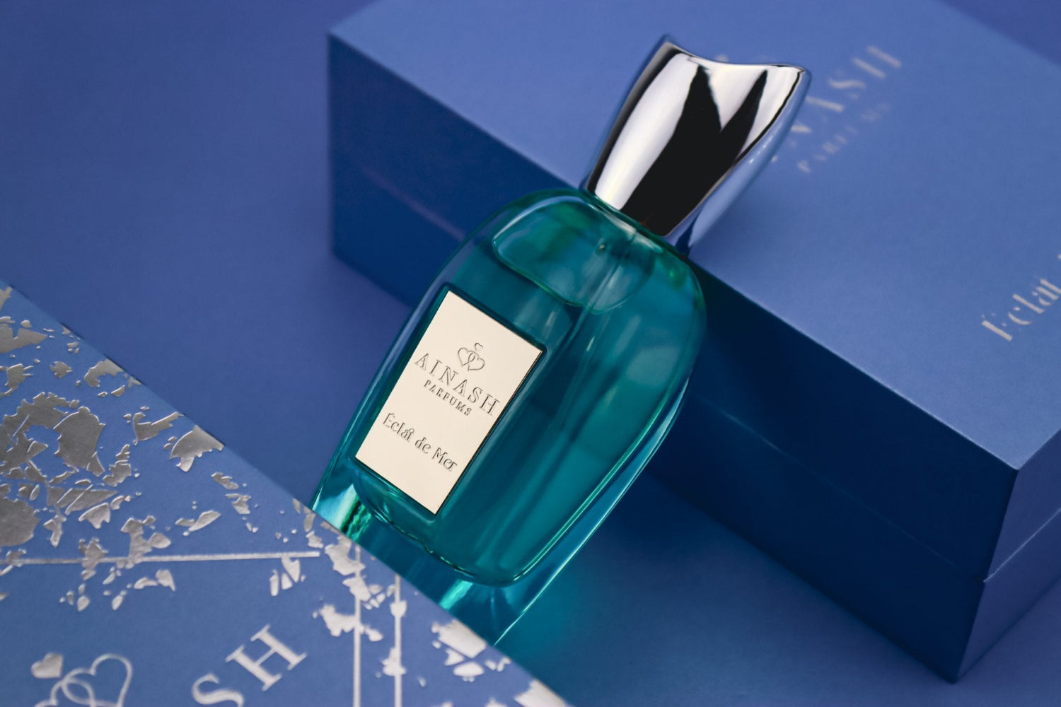 Teal perfume bottle of Éclat de Mer with silver cap placed on luxury blue box with metallic map details
