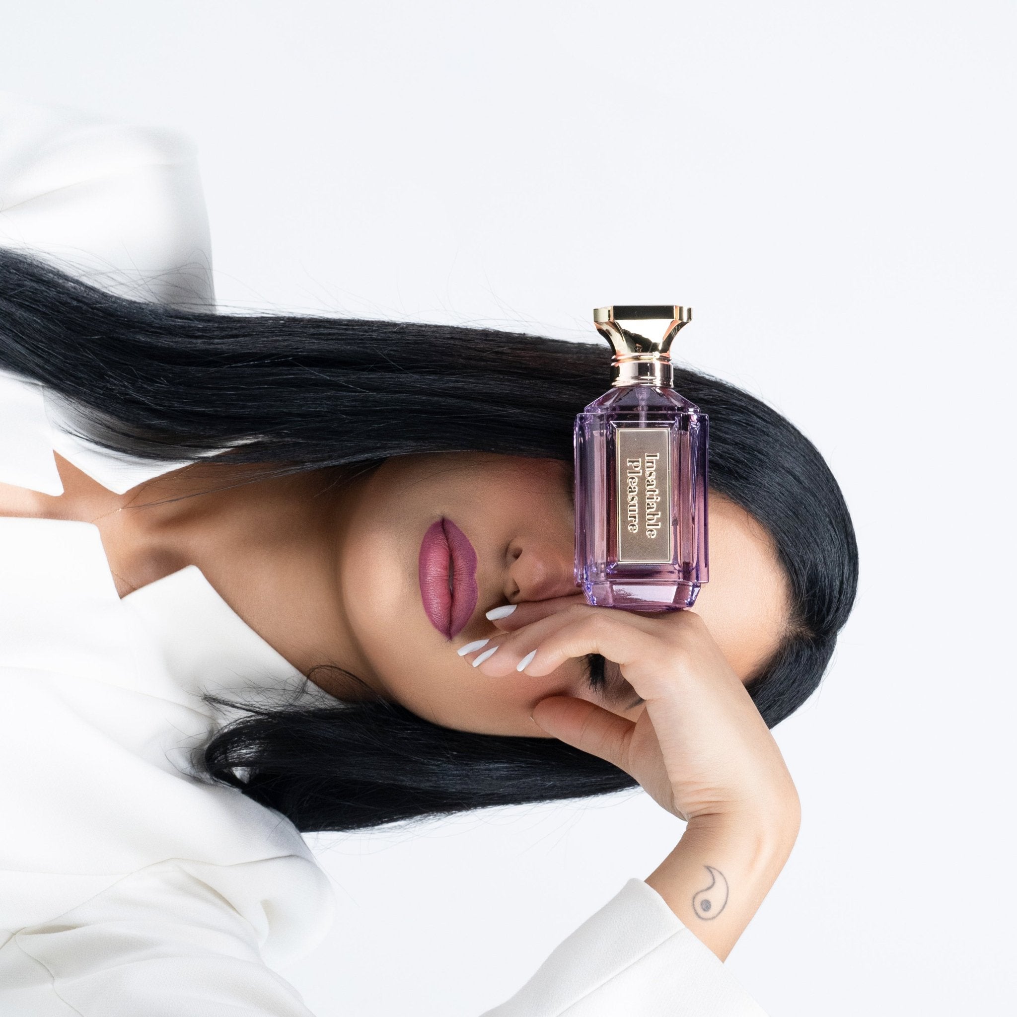 "Woman with long black hair holding Ainash Parfums’ Insatiable Pleasure — purple extrait de parfum bottle with gold plaque, posed against clean white background"
